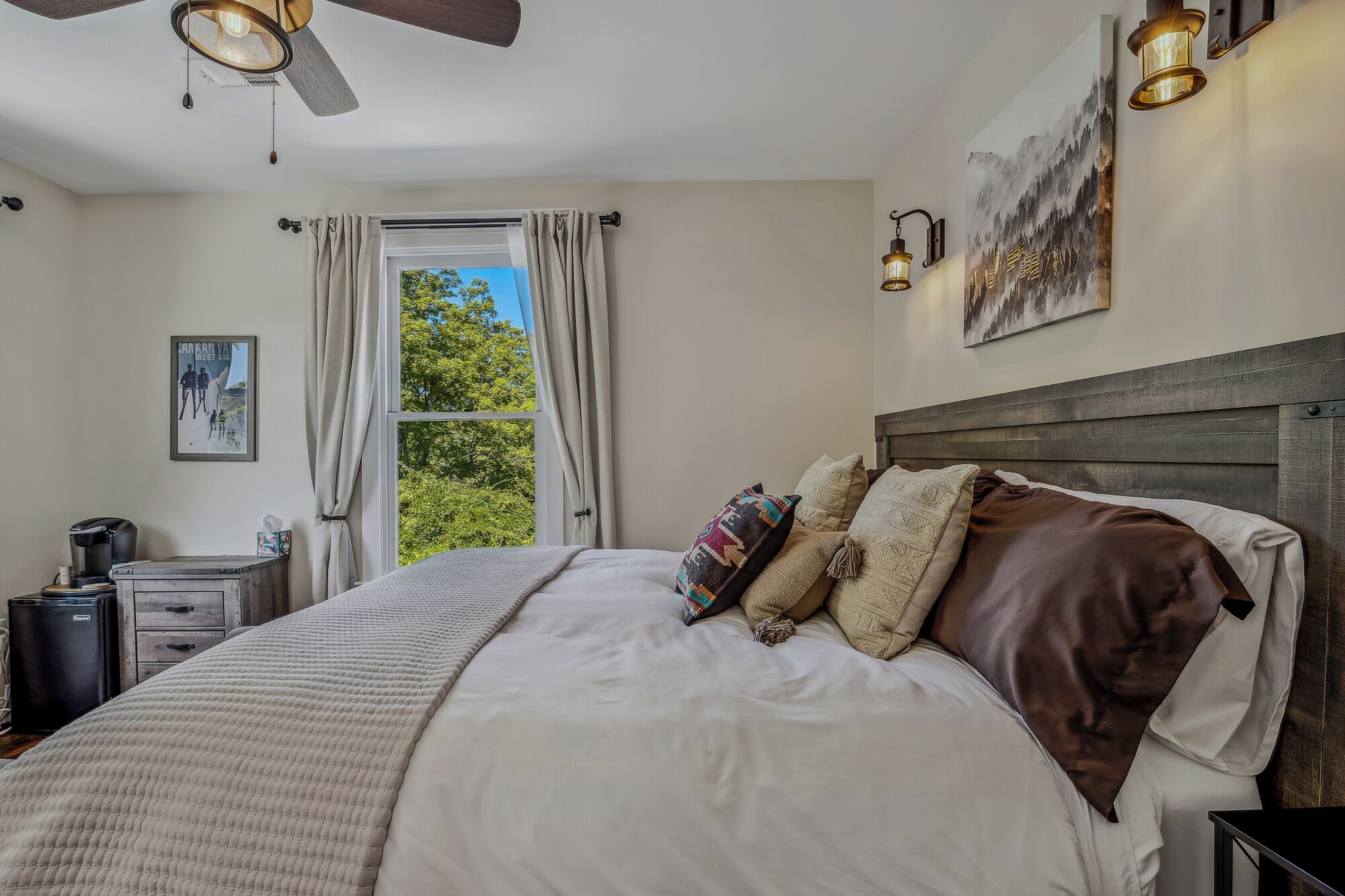 A bedroom with a large bed, a ceiling fan, a window with curtains, a bedside table, a lamp, and a painting on the wall.