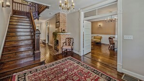A spacious hallway with a wooden staircase, a brick fireplace, and a dining area with a yellow chair. - Appalachian Suite In Town!- The Grand Castalian Inn B&B (Berkeley Springs)