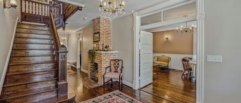 A spacious hallway with a wooden staircase, a brick fireplace, and a dining area with a yellow chair.