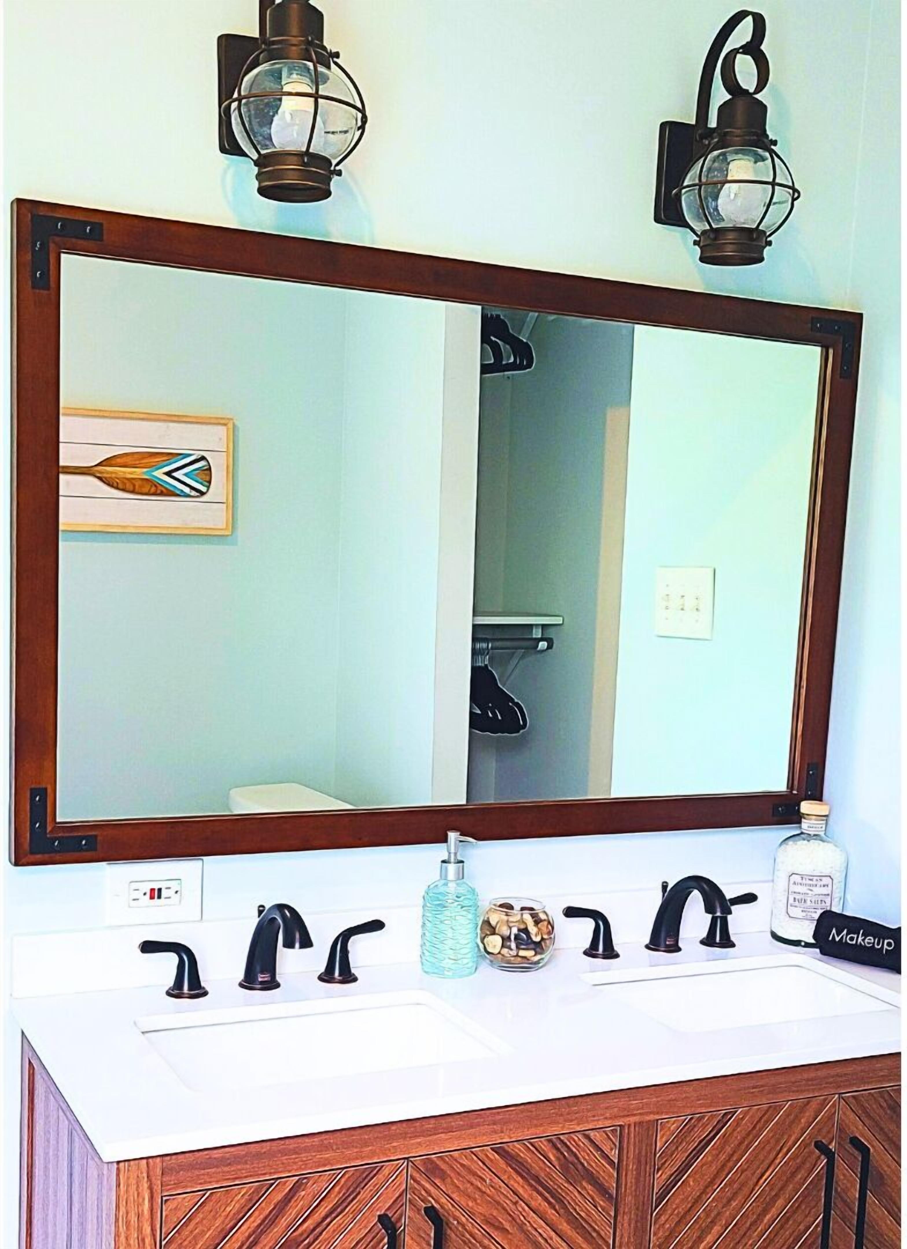 Une salle de bain avec un grand miroir, deux appliques murales, un lavabo avec des robinets noirs, un distributeur de savon et une bouteille d’alcool.