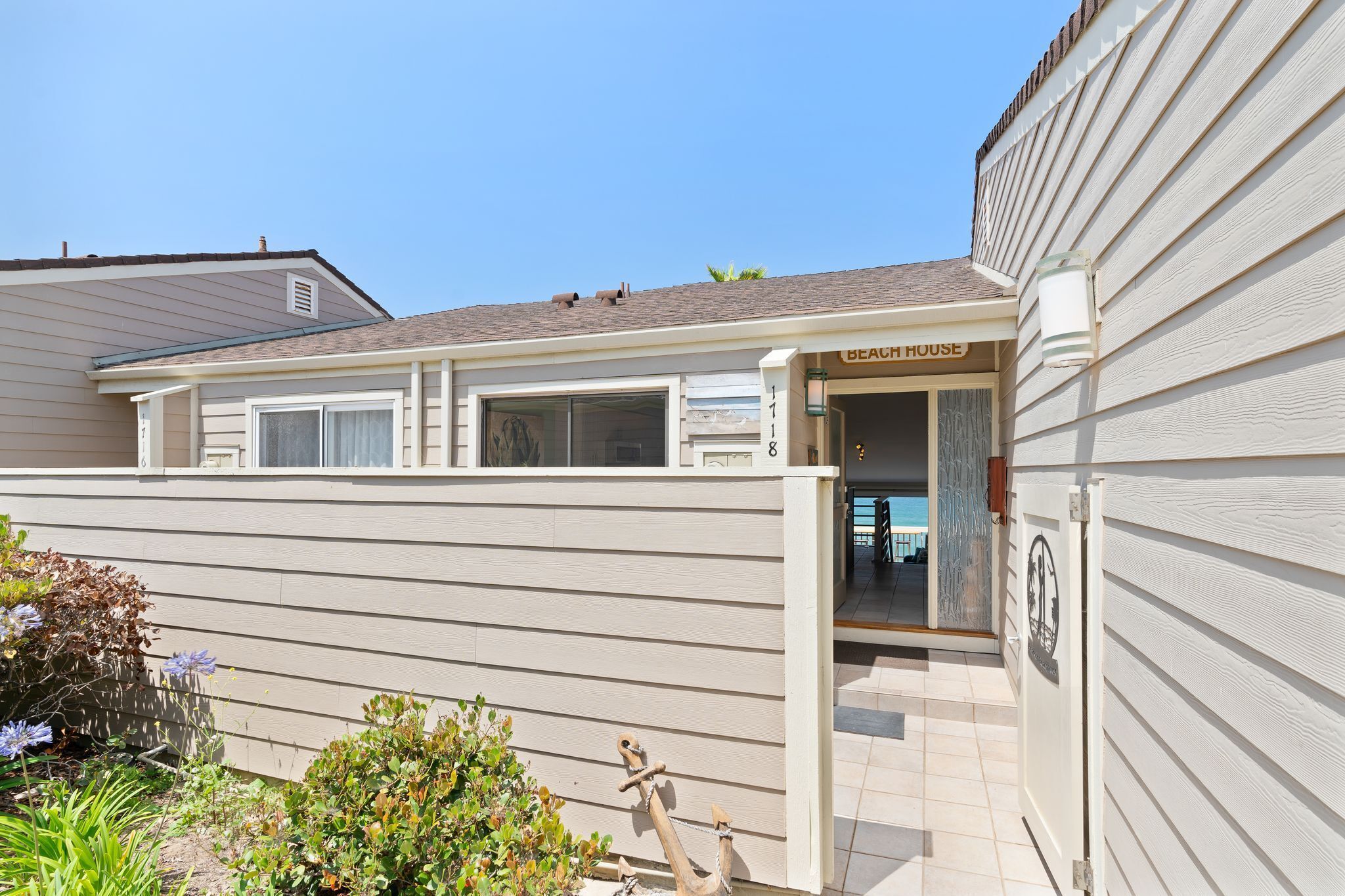 Ein zweigeschossiges Haus mit einem 'Beach House'-Schild, einer überdachten Terrasse und einem kleinen Garten mit Pflanzen und Blumen.