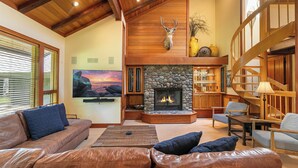 A spacious living room with a stone fireplace, leather sofas, a television, and a wooden staircase. - Chambers Lakeside Retreat - Walk to Lake, Views & HOA Gym, Pool + Beach (Homewood)