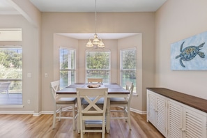 A dining area with a wooden table and chairs, a pendant light, and a view of greenery through the windows.