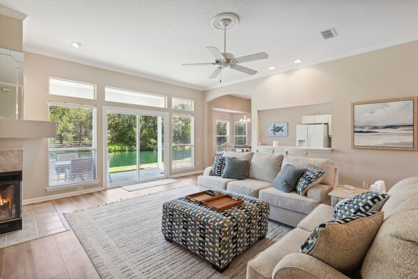 A spacious living room with a fireplace, large windows, and a ceiling fan.