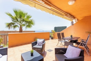 A rooftop terrace with outdoor furniture, a palm tree, and a view of the sea.