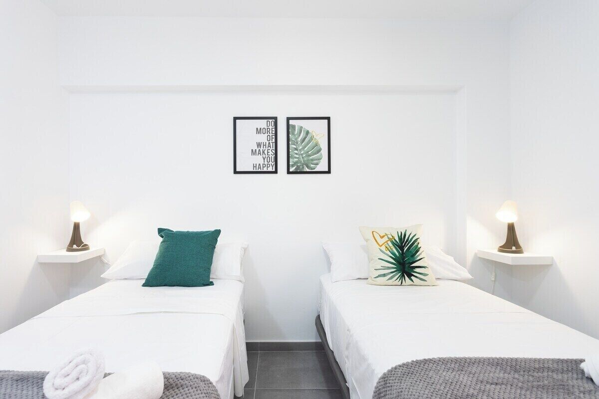 Una habitación de hotel con dos camas, ropa de cama blanca, una almohada verde y una almohada con forma de corazón, además de una cita motivacional enmarcada en la pared.