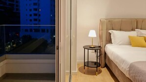 Habitación de hotel con cama, mesita de noche con lámpara, ventana con vistas a edificios y un equipo de aire acondicionado en la pared.