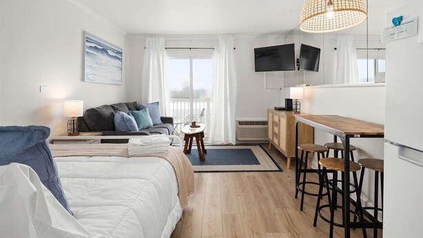 Een moderne hotelkamer met een bed, een bank, een eethoek met krukken, een televisie en een groot raam met gordijnen.