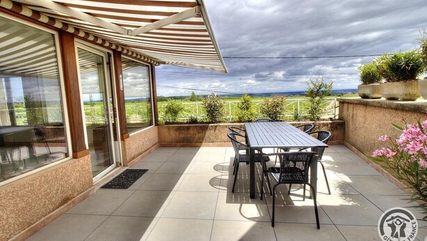 Eine überdachte Terrasse mit Tisch und Stühlen, großen Fenstern und Blick auf die Landschaft.