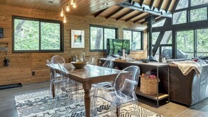 A modern cabin interior with a wooden dining table, clear chairs, and a sofa. Large windows offer views of the surrounding forest. - Lakefront Chalet w/ Fire Pit (Innsbrook)