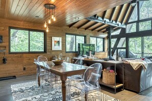 L’intérieur de ce chalet moderne est doté d’une table à manger en bois, de chaises transparentes et d’un canapé. De grandes fenêtres offrent une vue sur la forêt environnante.