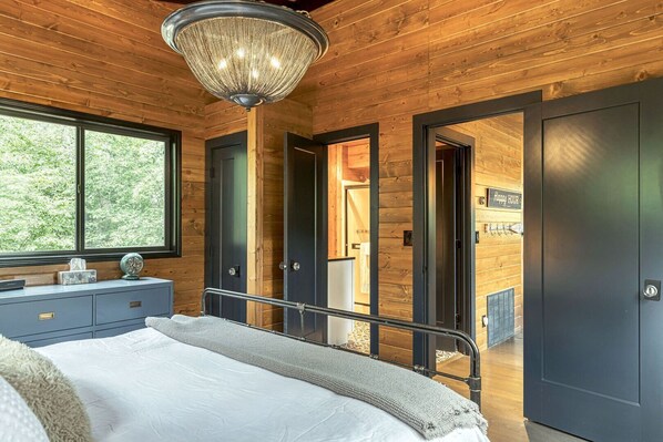 A bedroom with a bed, bedside table, and a large window. There is a chandelier hanging from the ceiling and a door leading to another room. - Lakefront Chalet w/ Fire Pit (Innsbrook)