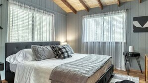 Une chambre à coucher avec un lit, deux fenêtres avec des rideaux légers, un ventilateur de plafond et une petite table de chevet.