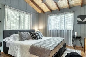 A bedroom with a bed, two windows with sheer curtains, a ceiling fan, and a small side table.