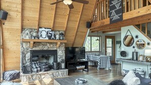Un salon chaleureux avec une cheminée en pierre, des poutres apparentes, un ventilateur de plafond et une nappe à carreaux.
