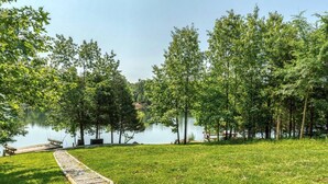 Un área verde con un camino de madera que lleva a un muelle junto a un lago tranquilo, rodeado de árboles.