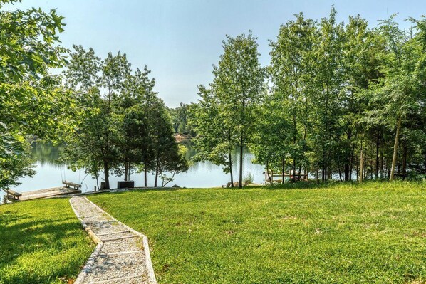 Un'area erbosa con un sentiero in legno che conduce a un molo su un lago calmo, circondato da alberi.