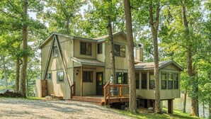 Une maison à deux étages avec une terrasse, entourée d’arbres et donnant sur une allée gravillonnée.