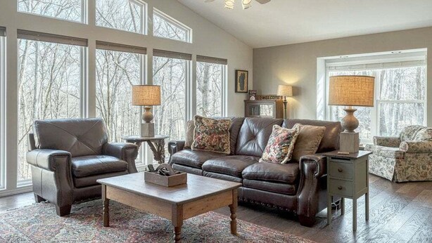 A spacious living room with a leather sofa, a wooden coffee table, and two armchairs. There are large windows providing natural light and a view of trees outside. - Innsbrook Home | Lake + Resort Access (Innsbrook)