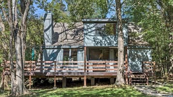 Escape to a secluded cabin surrounded by trees, featuring a chimney, large windows, and a wooden deck.