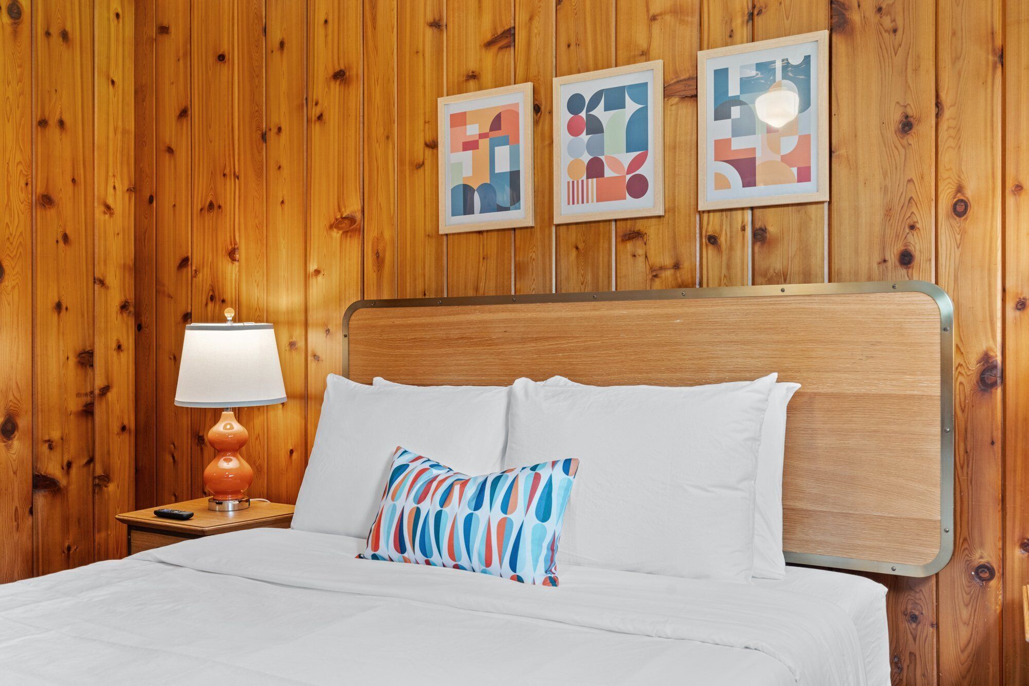 Une pièce avec un mur lambrissé en bois, un lit avec une literie blanche, un coussin coloré, une lampe sur une table de chevet en bois et trois œuvres d’art abstraites accrochées au mur.