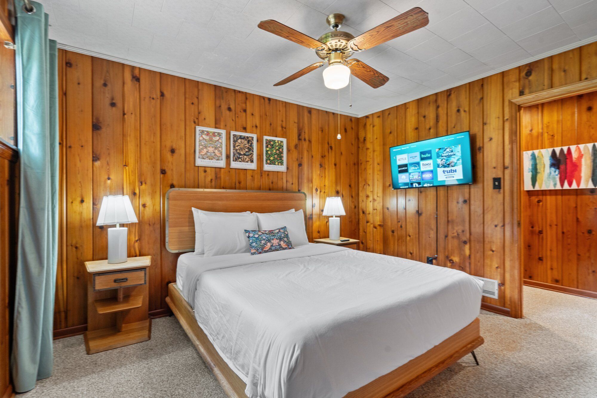 A wooden-paneled bedroom with a bed, two bedside tables with lamps, a ceiling fan, and a TV on the wall.