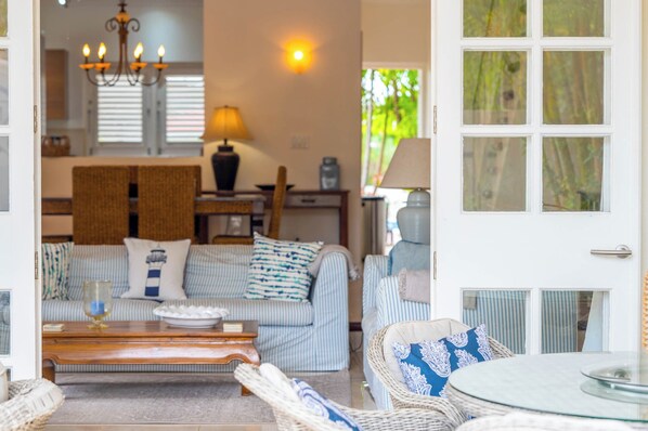 Un salon chaleureux avec un canapé, une table basse et un fauteuil en osier. On aperçoit une table à manger avec des chaises au fond.