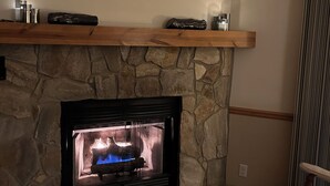A stone fireplace with a modern design, a wooden mantel shelf, and a comfortable seating area with a chair. - Beautiful Misty Mountain Views from a quaint cottage in Maggie Valley! (Maggie Valley)