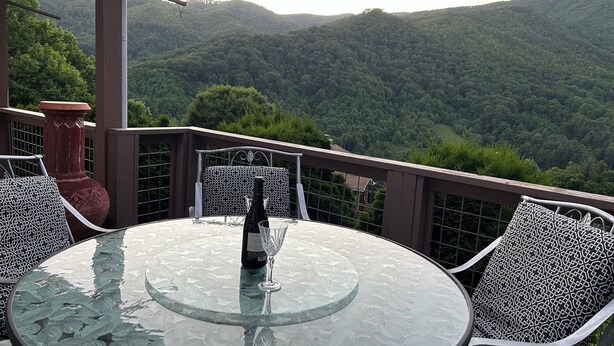 A round glass table with a bottle and glass on it, surrounded by patterned chairs, set on a balcony with a view of a forested mountain range. - Beautiful Misty Mountain Views from a quaint cottage in Maggie Valley! (Maggie Valley)