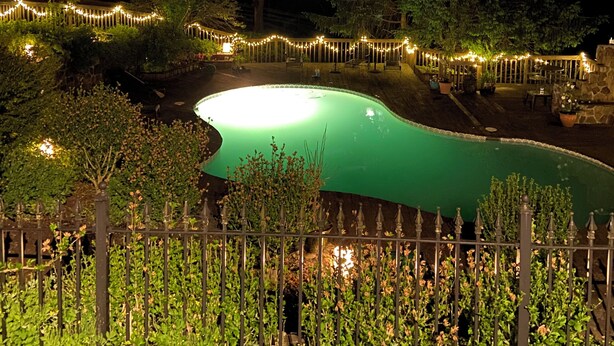 A nighttime view of a swimming pool surrounded by trees and a fence with string lights. - Designer Decorated Estate in Historic Downtown Lewisburg, West Virginia (Lewisburg)