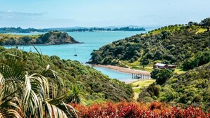 A coastal landscape with a beach, a small house, and lush greenery. - Delamore Cove Kāraka Studio (Waiheke Island)