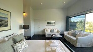 A modern hotel room with a large bed, two seating areas, a desk, and a large window with a view of greenery. - Delamore Cove Kāraka Studio (Waiheke Island)