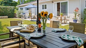 Un área de comedor al aire libre con una mesa preparada para dos, que incluye una botella de aceite de oliva, un jarrón con girasoles y una cesta de pan.