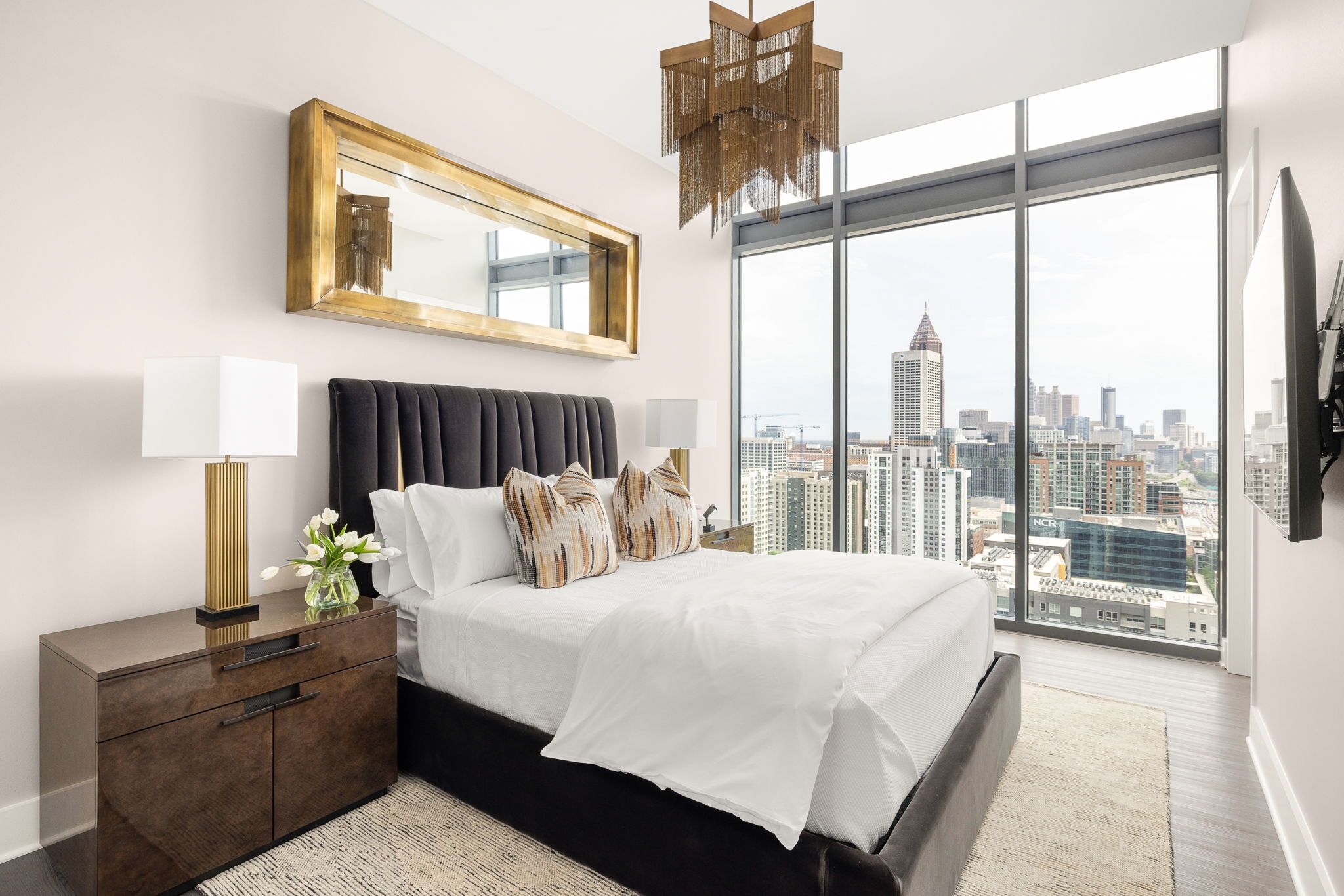 A modern bedroom with a large bed, a nightstand, a mirror, and a view of a city skyline.