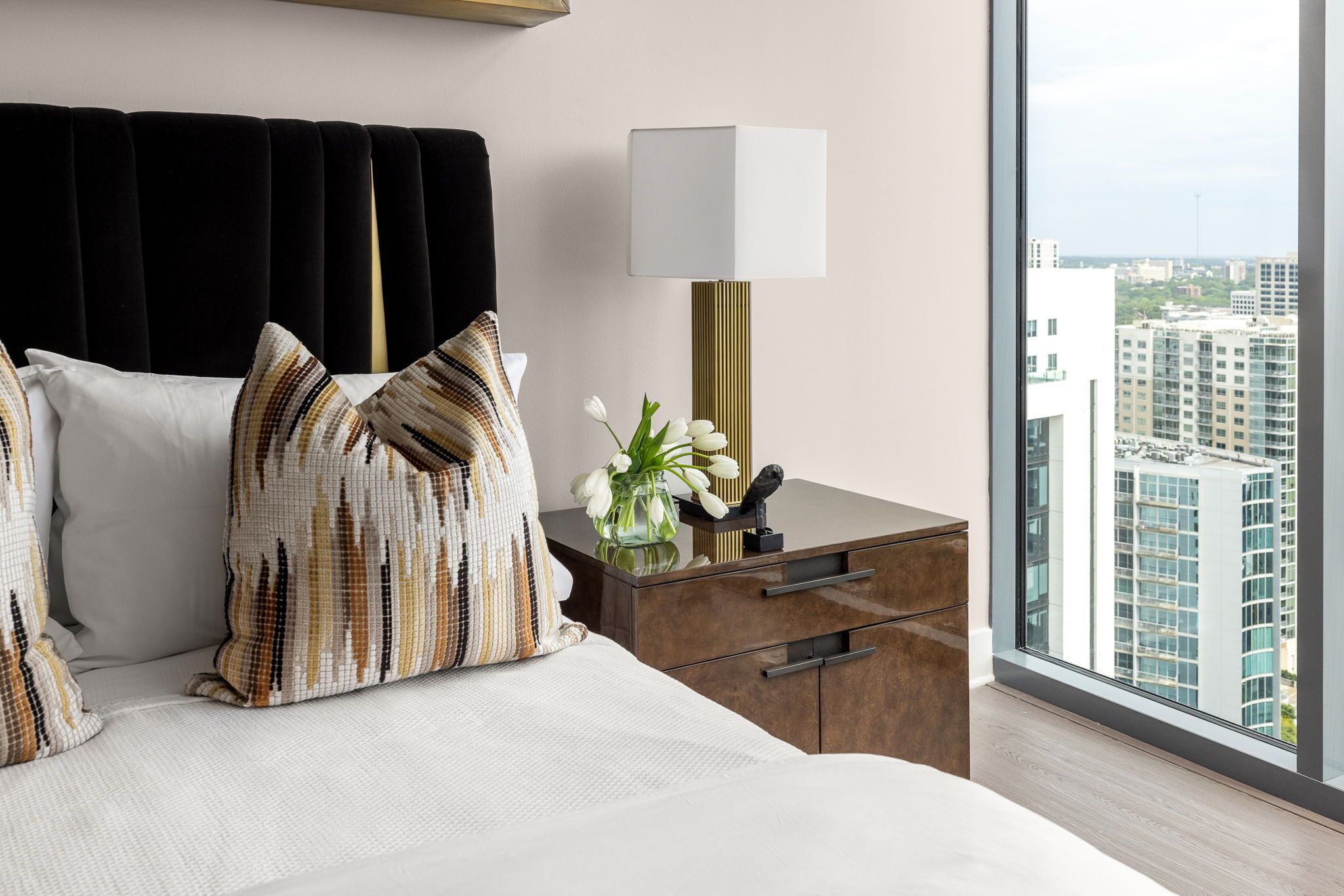 Quarto de hotel moderno com uma cama grande, uma mesa de cabeceira, uma luminária e vista para a cidade.