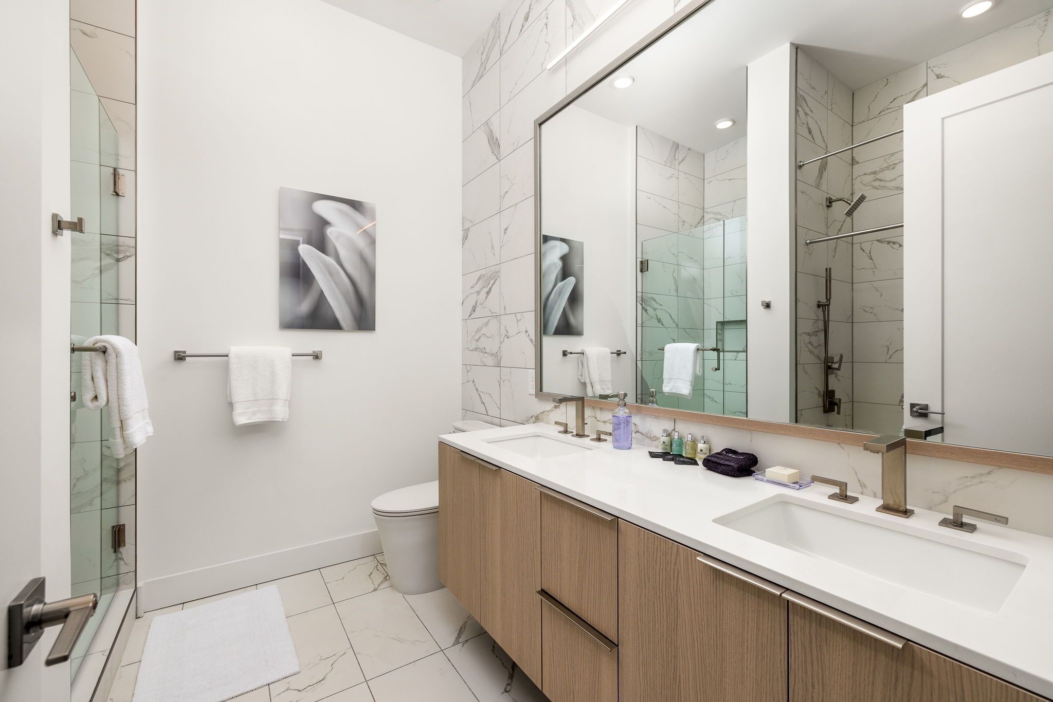 Un bagno moderno con pavimento in marmo, uno specchio grande, un lavandino bianco e mobili in legno.