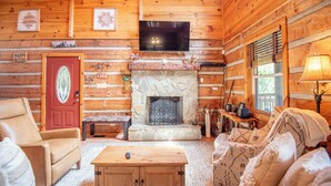 A cozy log cabin interior with a stone fireplace, a television mounted above it, and a comfortable seating area with a sofa and armchairs. - Bear Haven | Overlooks a Bass-Stocked Pond | Pool Table & Card Table (Grassy Creek)