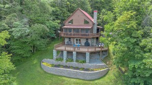 A two-story cabin with a stone foundation, a red roof, and a deck with outdoor seating. - Bear Haven | Overlooks a Bass-Stocked Pond | Pool Table & Card Table (Grassy Creek)