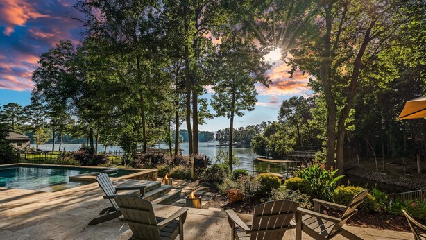 An outdoor patio with Adirondack chairs, a fire pit, and a view of a lake with a bridge in the background. - Lake Effect by SoCharm | Pool & Spa, Waterfront (Mooresville)
