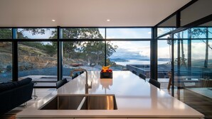 A modern kitchen with a large island, stainless steel sink, and a view of a rocky coastline and ocean. - Mountain Top Snug Whole Property Itas (Snug)