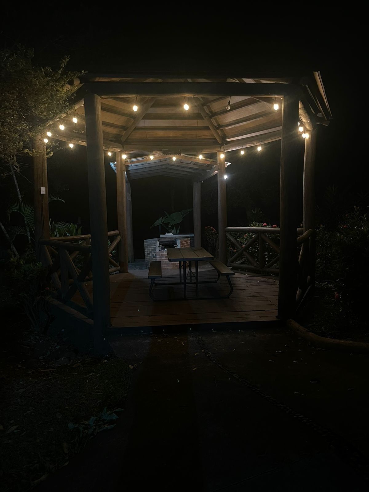Un pabellón de madera con luces colgantes, una mesa de picnic y una chimenea de ladrillo.