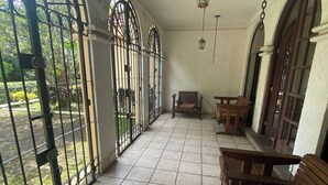 Un patio cubierto con piso de baldosas, bancos de madera y vista a un jardín a través de portones metálicos.