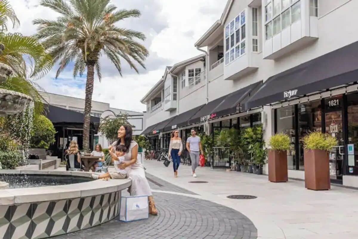 A public square with a fountain, palm trees, and modern buildings with storefronts.