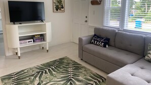 Een woonkamer met een grijze bank, een flatscreen televisie op een witte standaard, een boekenkast met gezelschapsspellen, een vloerkleed met een tropisch bladmotief en een raam met jaloezieën.