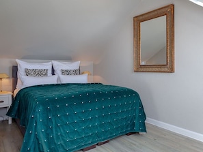 Un lit bien fait, recouvert d’une couvre-lit turquoise, avec deux oreillers blancs aux housses à motifs, et un miroir à cadre doré accroché au mur.