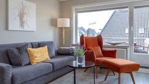 A living room with a grey sofa, an orange armchair, a small table with a plant, and a view of a cityscape through large windows. - Strand Brise - "Strand-Brise" vacation apartment in Westerland, in a prime location (Sylt)