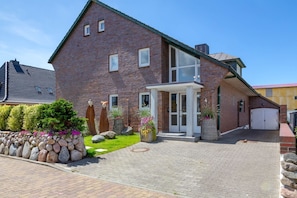 Une maison à deux étages, construite en briques, avec un porche à l’avant, un garage et un jardin bien entretenu agrémenté de rochers et de plantes.