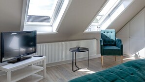 A modern loft bedroom with skylights, a flat-screen TV, a comfortable teal armchair, and a tufted green sofa. - Strand Brise - "Strand-Brise" vacation apartment in Westerland, in a prime location (Sylt)