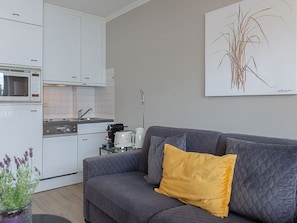 Une cuisine moderne avec des meubles blancs, un four à micro-ondes et une machine à café. On y trouve un canapé gris avec un coussin jaune et un tableau représentant de l’herbe accroché au mur.