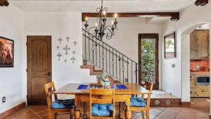 Un comedor con una mesa y sillas de madera, una escalera con barandilla de hierro forjado y una cocina visible a través de un arco.